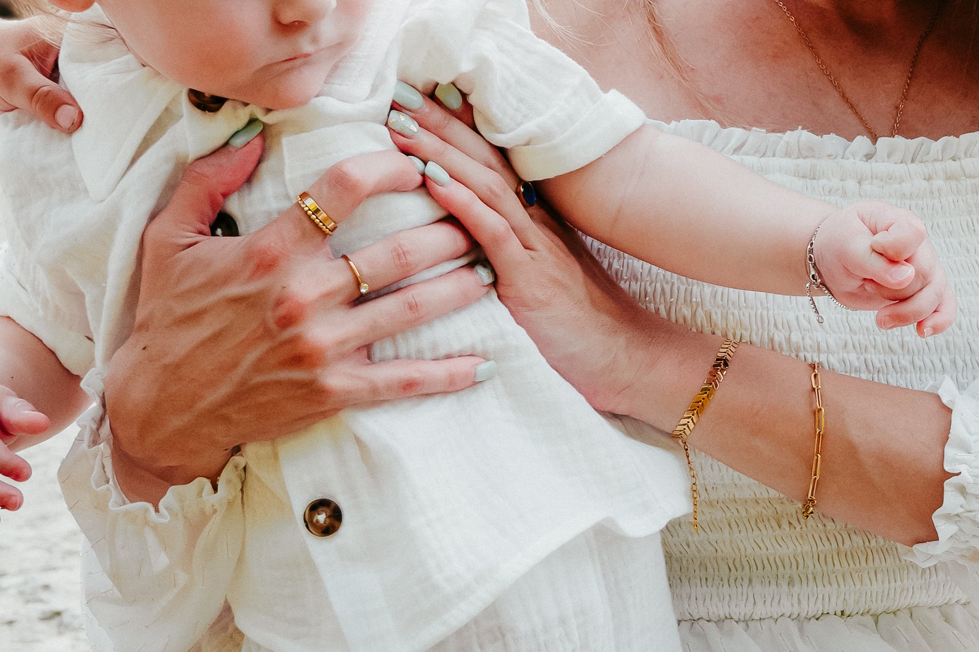 Baby in a white outfit held by an adult, with visible jewelry on hands.